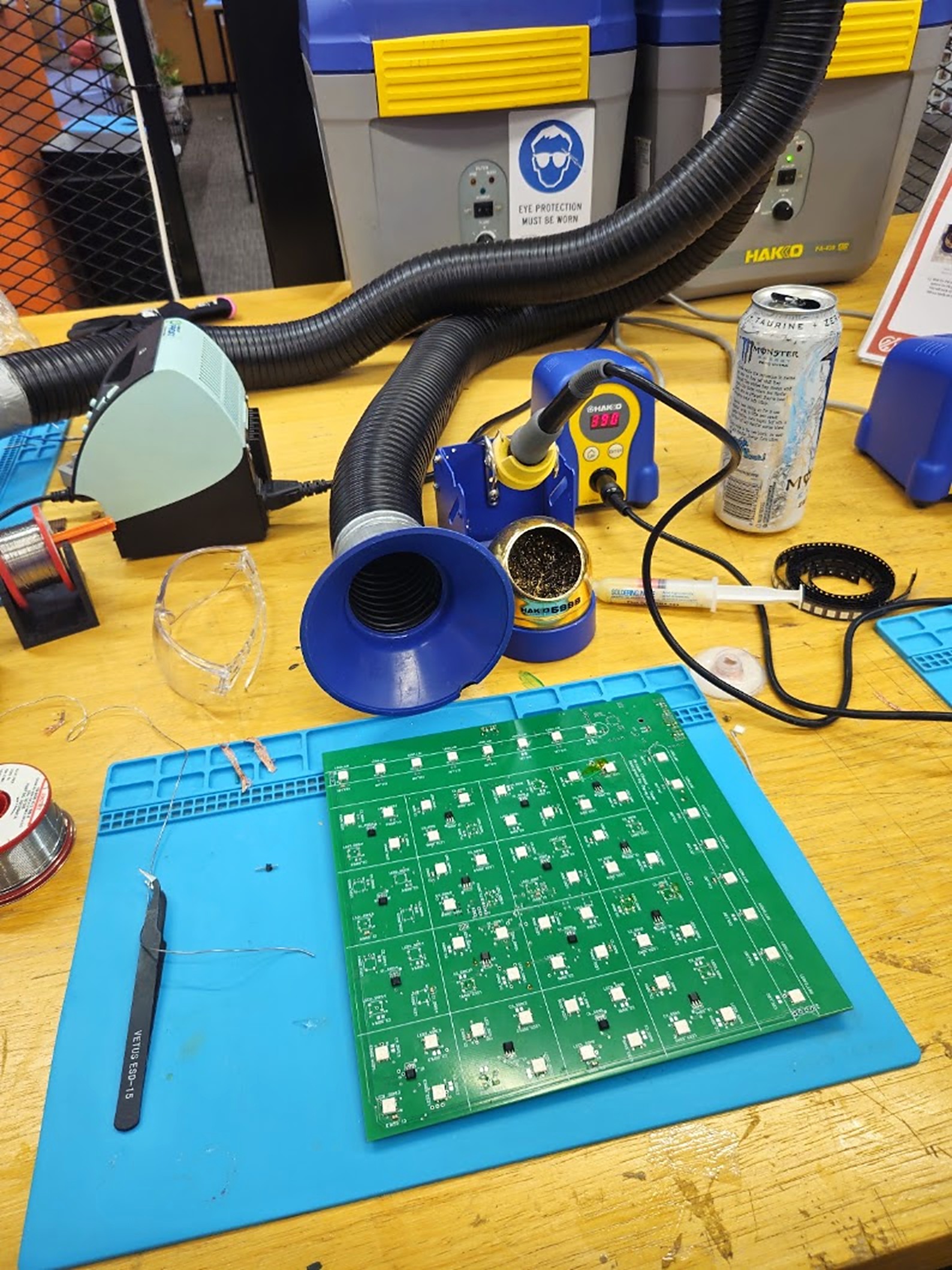 The PCB on a silicone mat at a hackathon soldering station, with fume extractors
