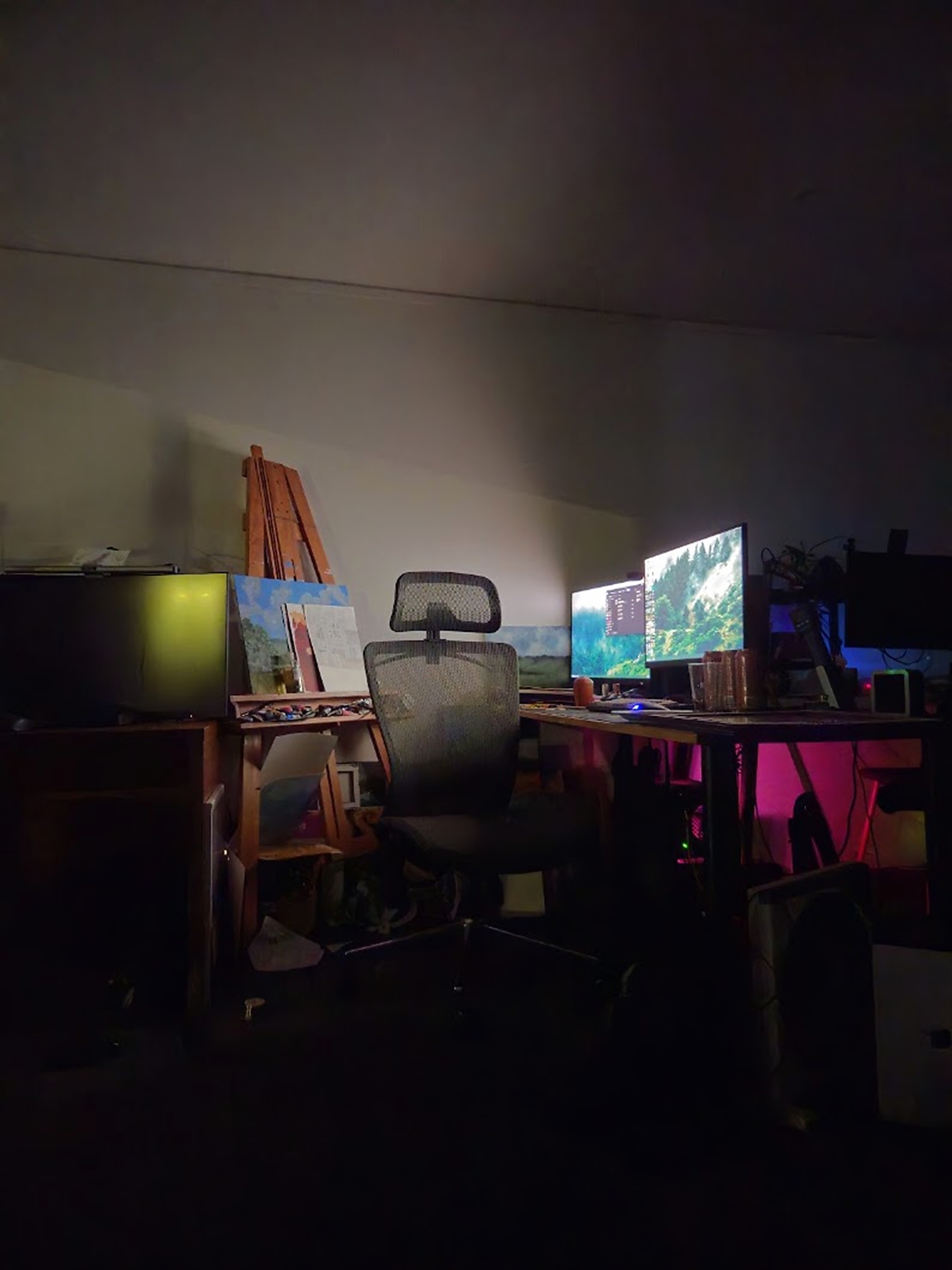 Ara's desk setup after dark: dual monitors glowing, pink light under the desk, and the easel beside the chair